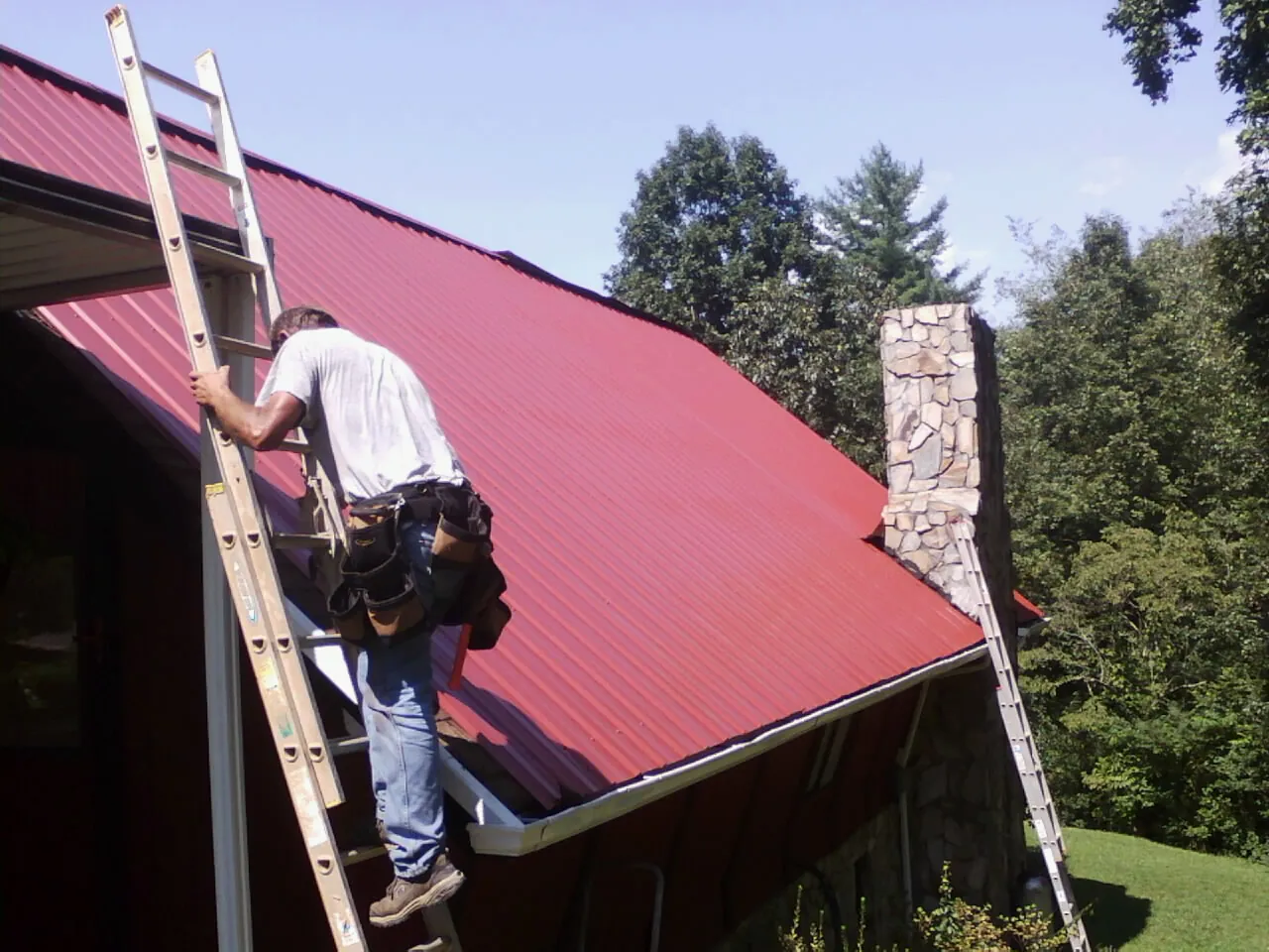 Roofer on a ladder beside a red metal roof and stone chimney — Soffit & Fascia Repair by Roofing Brothers in Bend